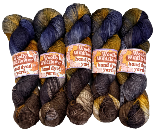 Five skeins of hand-dyed yarn in brown, blue, and orange on a white background with 'Woolly Wildflower' labels.