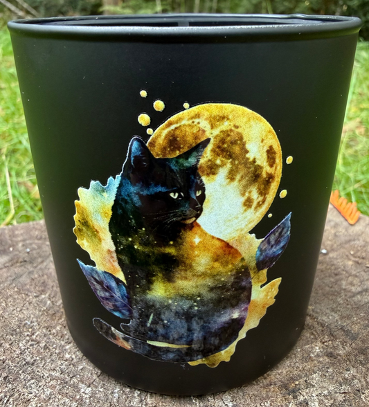 Black tumbler candle with a cat and moon design on a stone surface outdoors.