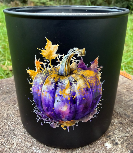 Black tumbler candle with a purple pumpkin design on a stone surface outdoors.
