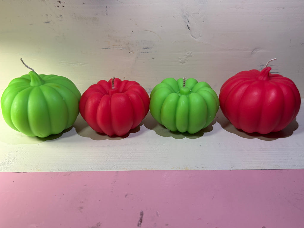 Four decorative pumpkin-shaped candles in alternating bright green and red, lined up on a white surface against a light wall.