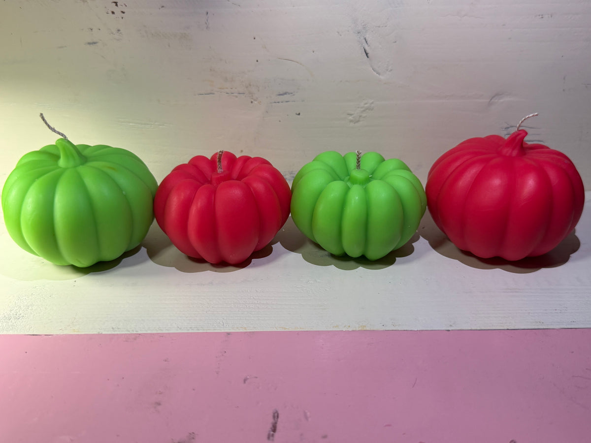 Four decorative pumpkin-shaped candles in alternating bright green and red, lined up on a white surface against a light wall.