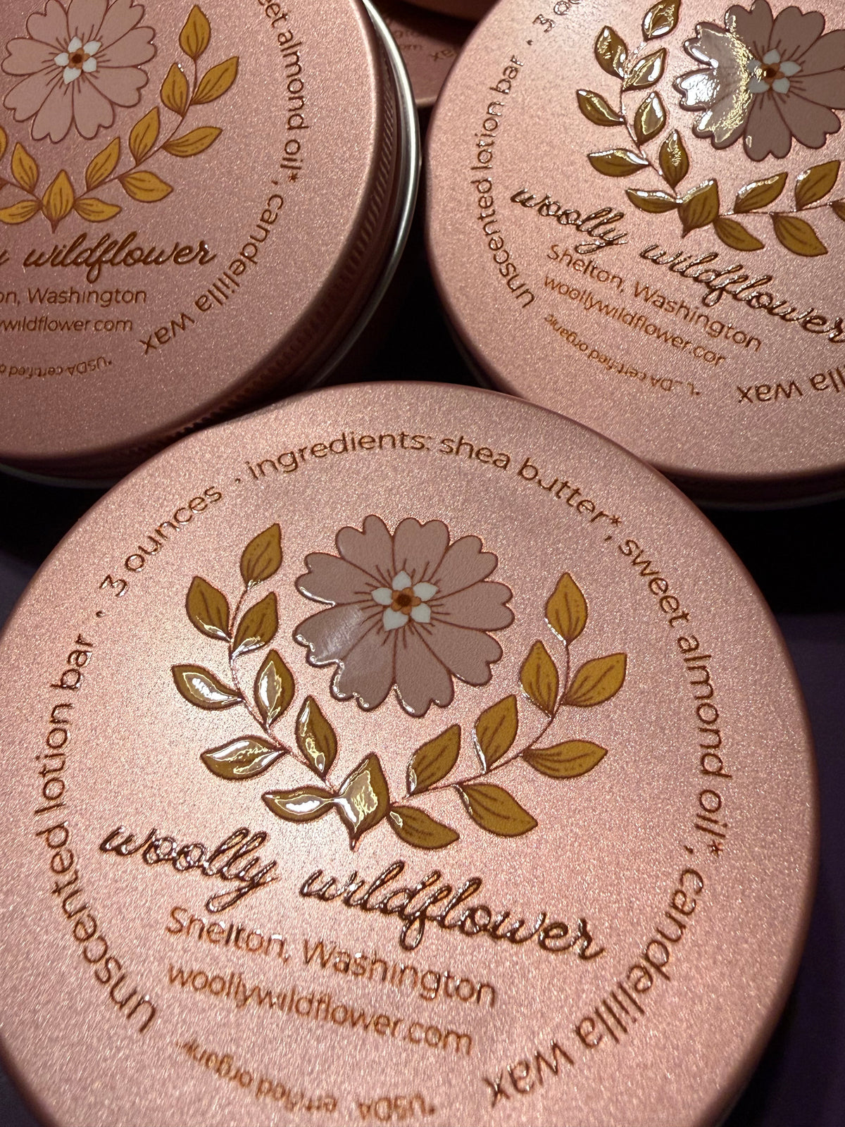 Three round metal tins of Woolly Wildflower unscented lotion bar, showing the rose gold lids with gold floral and leaf designs and product text.