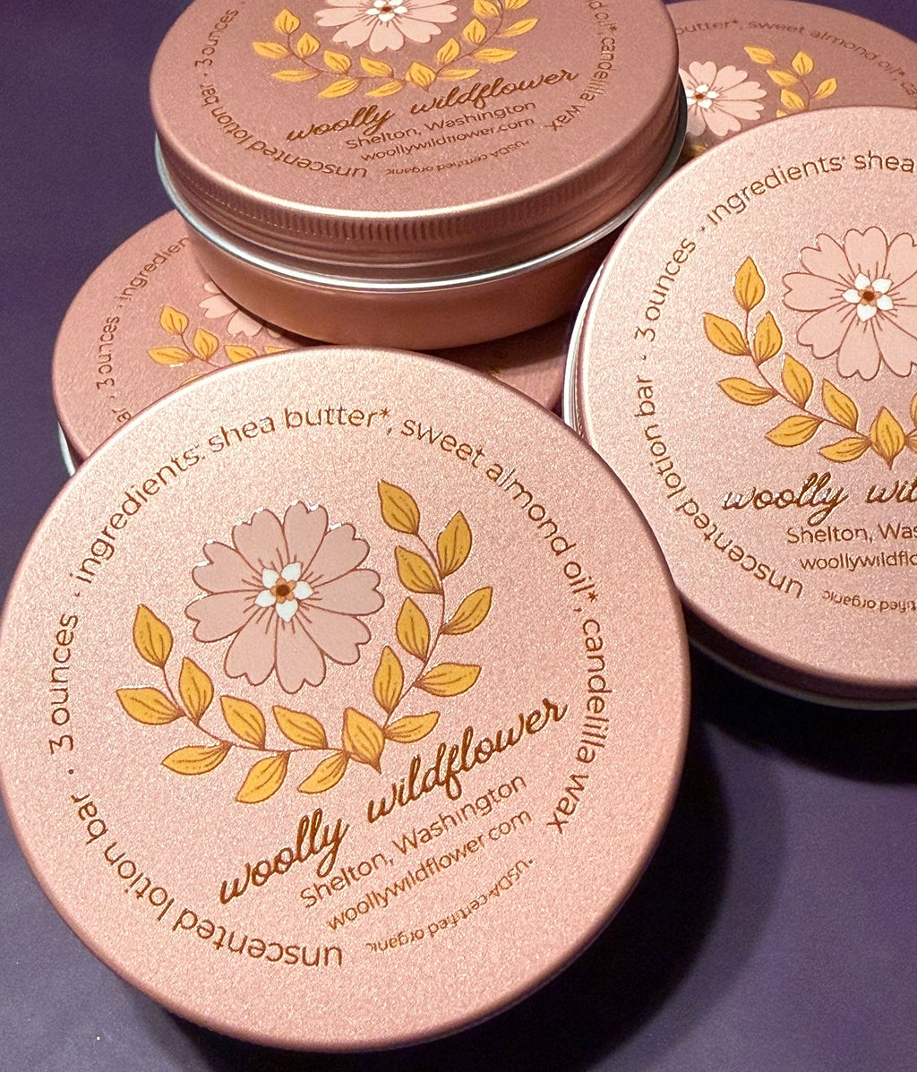 Several round metal tins of Woolly Wildflower unscented lotion bar are arranged on a dark purple surface. The lids are a warm metallic pink/rose tone with a floral illustration and gold text reading 'woolly wildflower' and listing ingredients such as shea butter, sweet almond oil, and camellia wax.