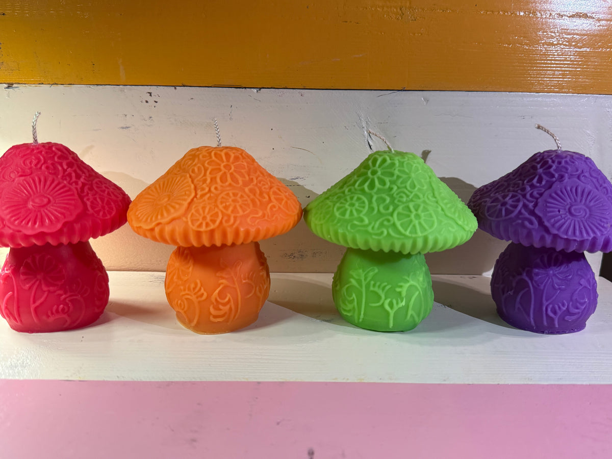 Four mushroom-shaped candles in pink, orange, green, and purple, arranged in a row on a white shelf against a colorful background.