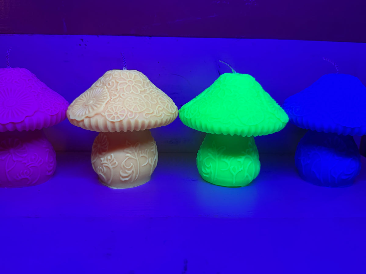 Four decorative mushroom-shaped candles in different colors—pink, cream, glowing neon green, and blue—arranged in a row under UV lighting that casts a strong blue and purple glow.