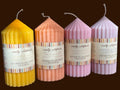 Four soy wax pillar candles in different pastel and bright colors are lined up on a wooden surface. The candles have ribbed sides, white wicks, and paper bands with the brand name 'wreatly wildflower' and scent names like maple sandalwood, pumpkin & cinnamon, jammy apple cider, and moonlight lavender.