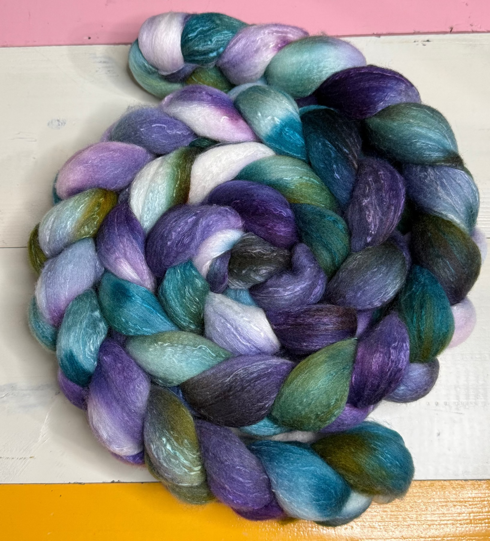 A coiled bundle of variegated wool roving or yarn with blended shades of purple, teal blue, green, and white, photographed on a painted surface with pink, white, and yellow sections visible.