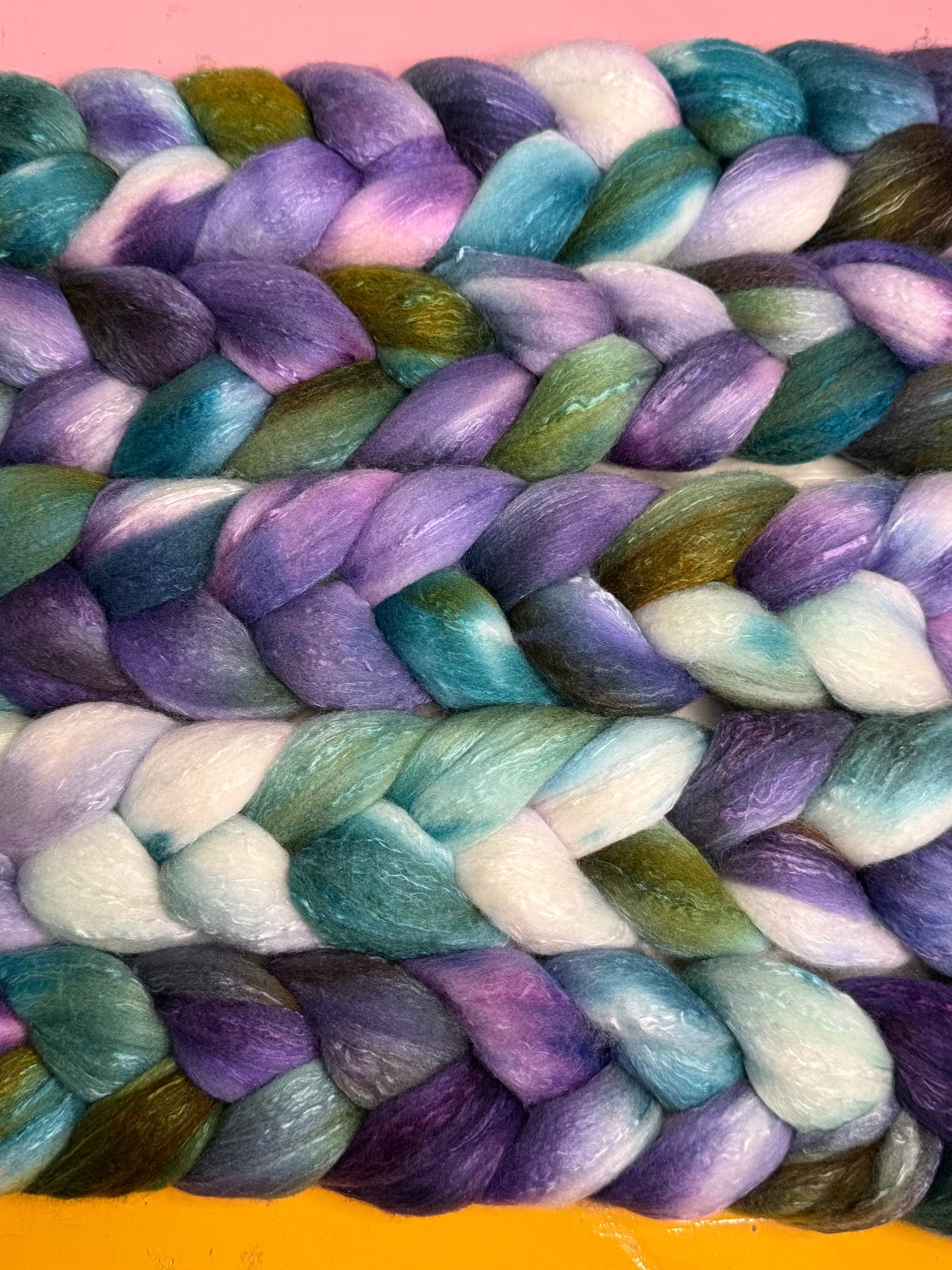 A close-up of a thick braided fiber wool roving, showing a variegated pattern of purple, teal blue, white, and olive green. The braid is photographed flat against a split background with a pink top section and a yellow-orange bottom section.