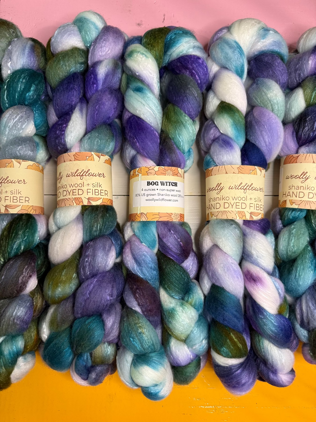 Five skeins of hand-dyed wool fiber yarn arranged side by side on a pink and yellow background. The yarn features variegated colors of purple, blue, teal, green, and white, with paper labels wrapped around the center.