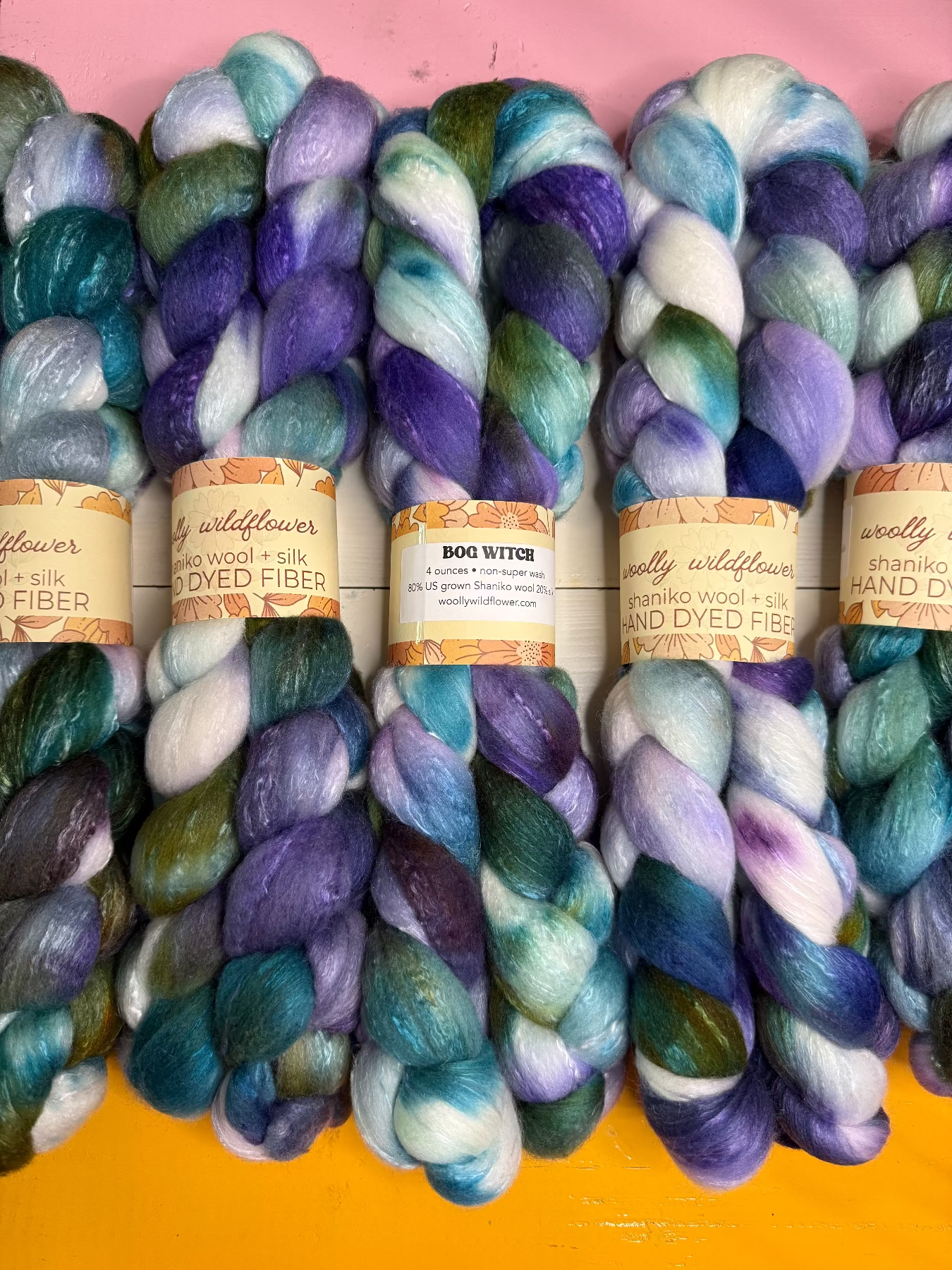 Five skeins of hand-dyed wool fiber yarn arranged side by side on a pink and yellow background. The yarn features variegated colors of purple, blue, teal, green, and white, with paper labels wrapped around the center.