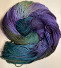 A skein of variegated yarn with a color-shifting gradient from purple and blue to teal and green, arranged in a spiral on a light off-white wooden background.