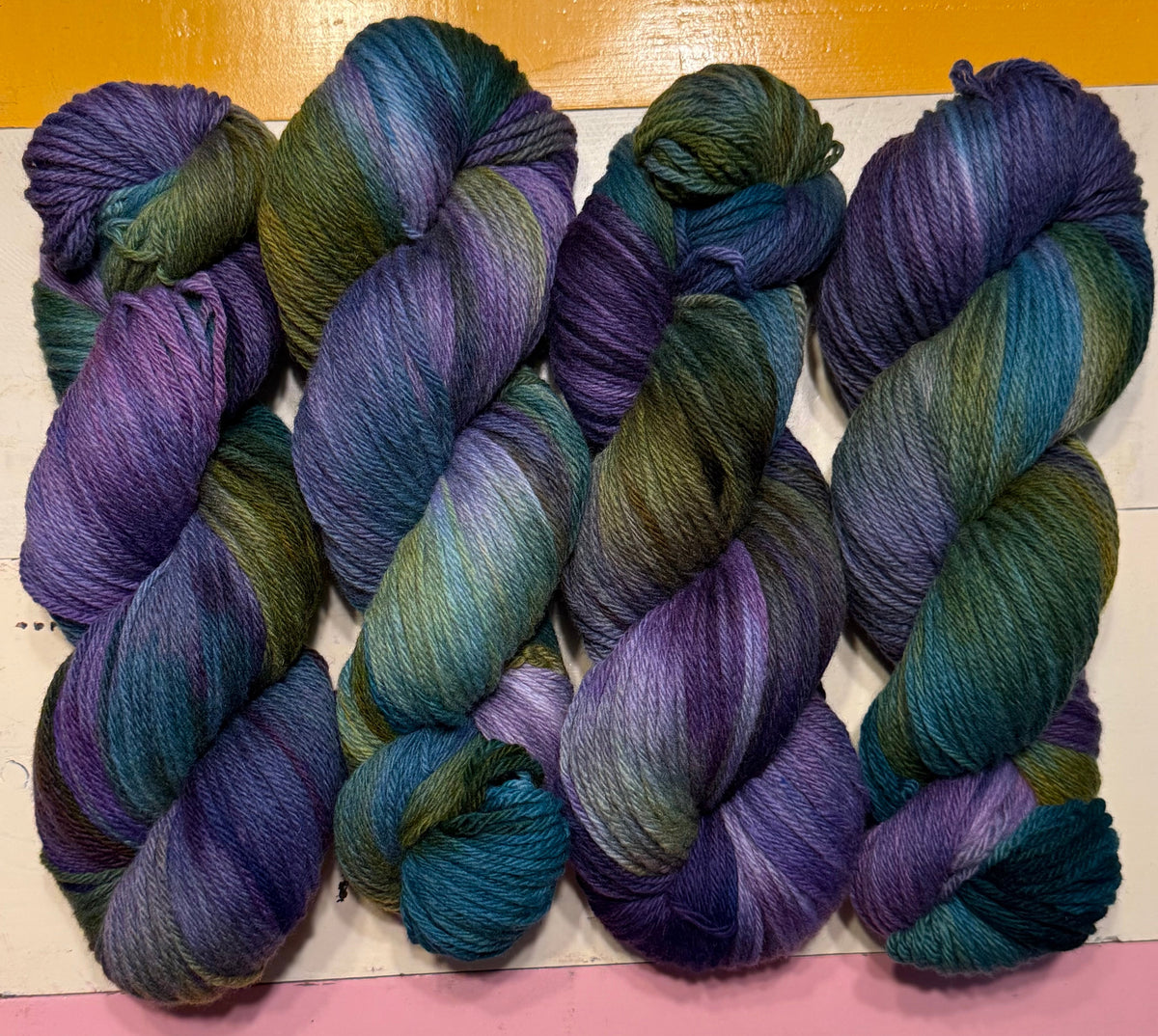 Six skeins of variegated yarn arranged side by side on a white surface, featuring color-shifting hues of purple, blue, teal, and olive green, set against a split yellow and pink background.