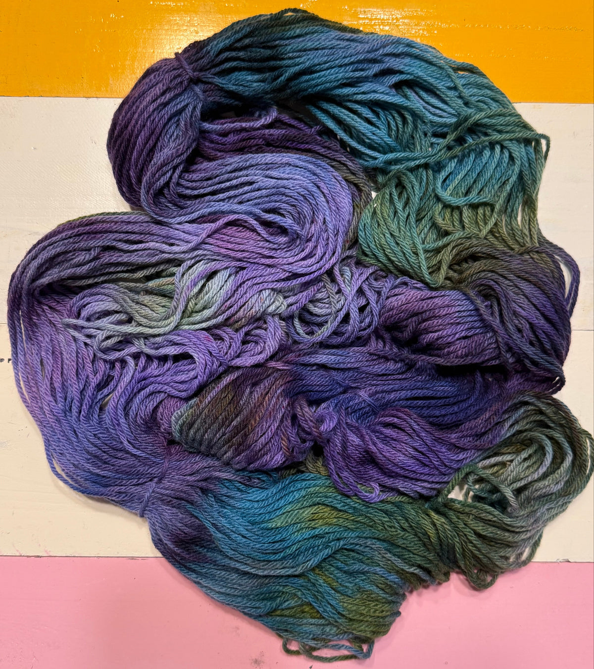 A pile of variegated yarn skeins showing a color-shifting gradient from purple and blue to teal and olive green, laid flat on a striped background of orange, white, and pink.