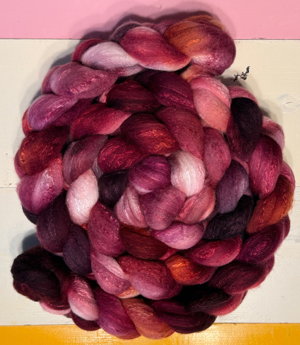 A coiled bundle of variegated wool roving in shades of deep magenta, burgundy, plum, and soft pink, displayed on a white surface with a yellow top section and a pink bottom section.