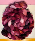 A coiled bundle of variegated wool roving in shades of deep magenta, burgundy, plum, and soft pink, displayed on a white surface with a yellow top section and a pink bottom section.