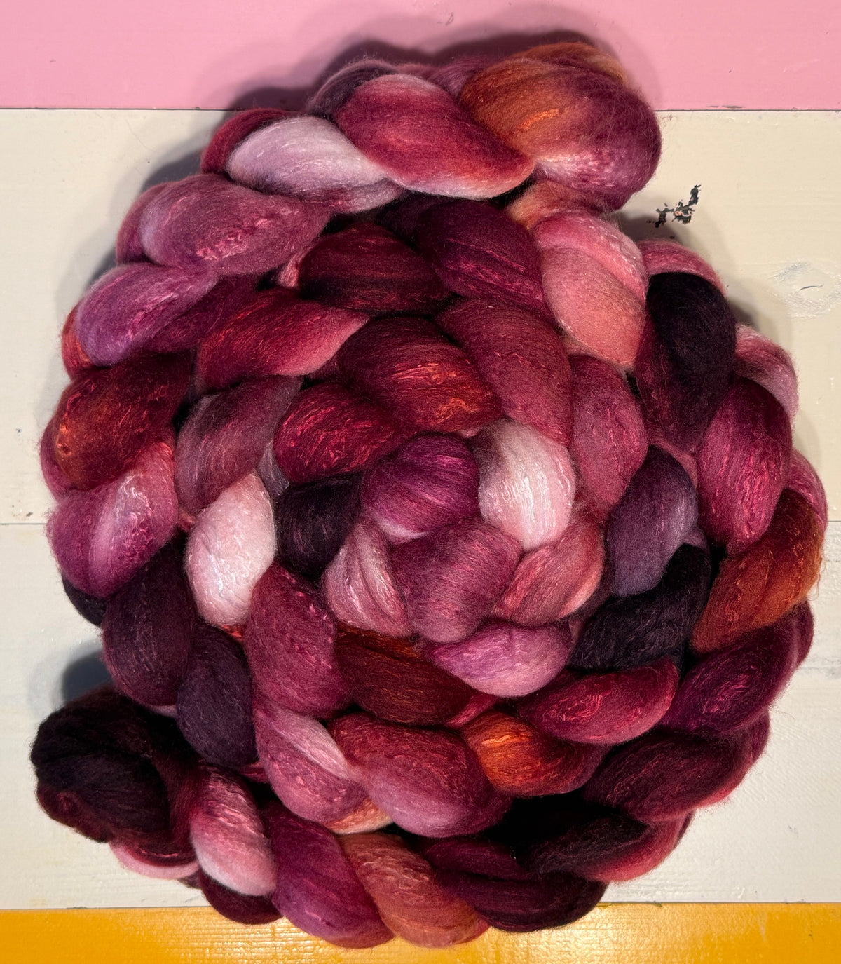 A coiled bundle of variegated wool roving in shades of deep magenta, burgundy, plum, and soft pink, displayed on a white surface with a yellow top section and a pink bottom section.
