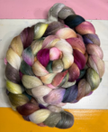A coiled bundle of soft, fuzzy yarn or fiber in mixed pastel and saturated colors, including pink, purple, cream, gray, green, and yellow, photographed against a pink, white, and yellow background.