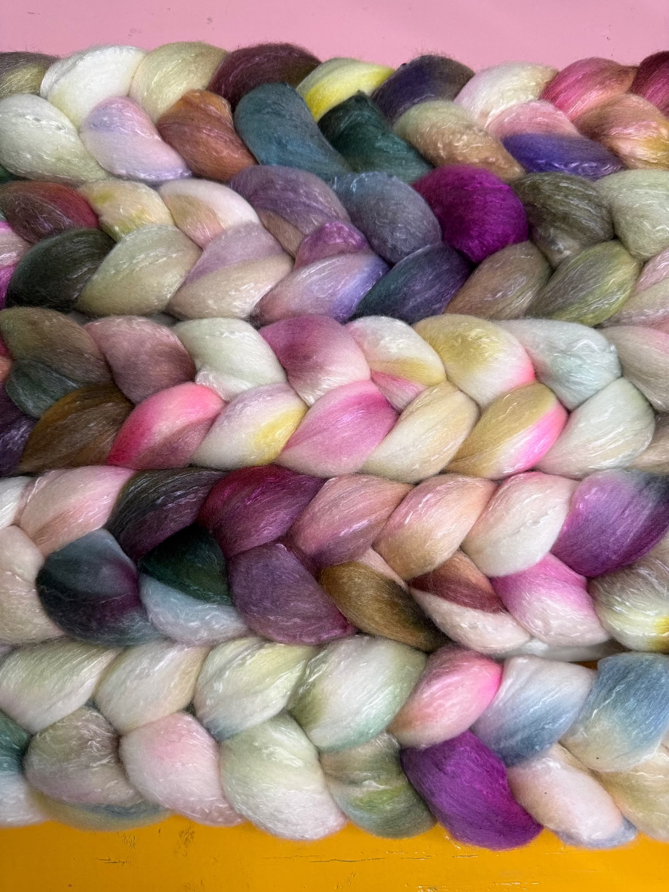 A close-up of a thick braided yarn or rope made from soft, fluffy fibers in many blended colors, including pink, purple, blue, green, yellow, and cream, arranged tightly against a pink and yellow background.