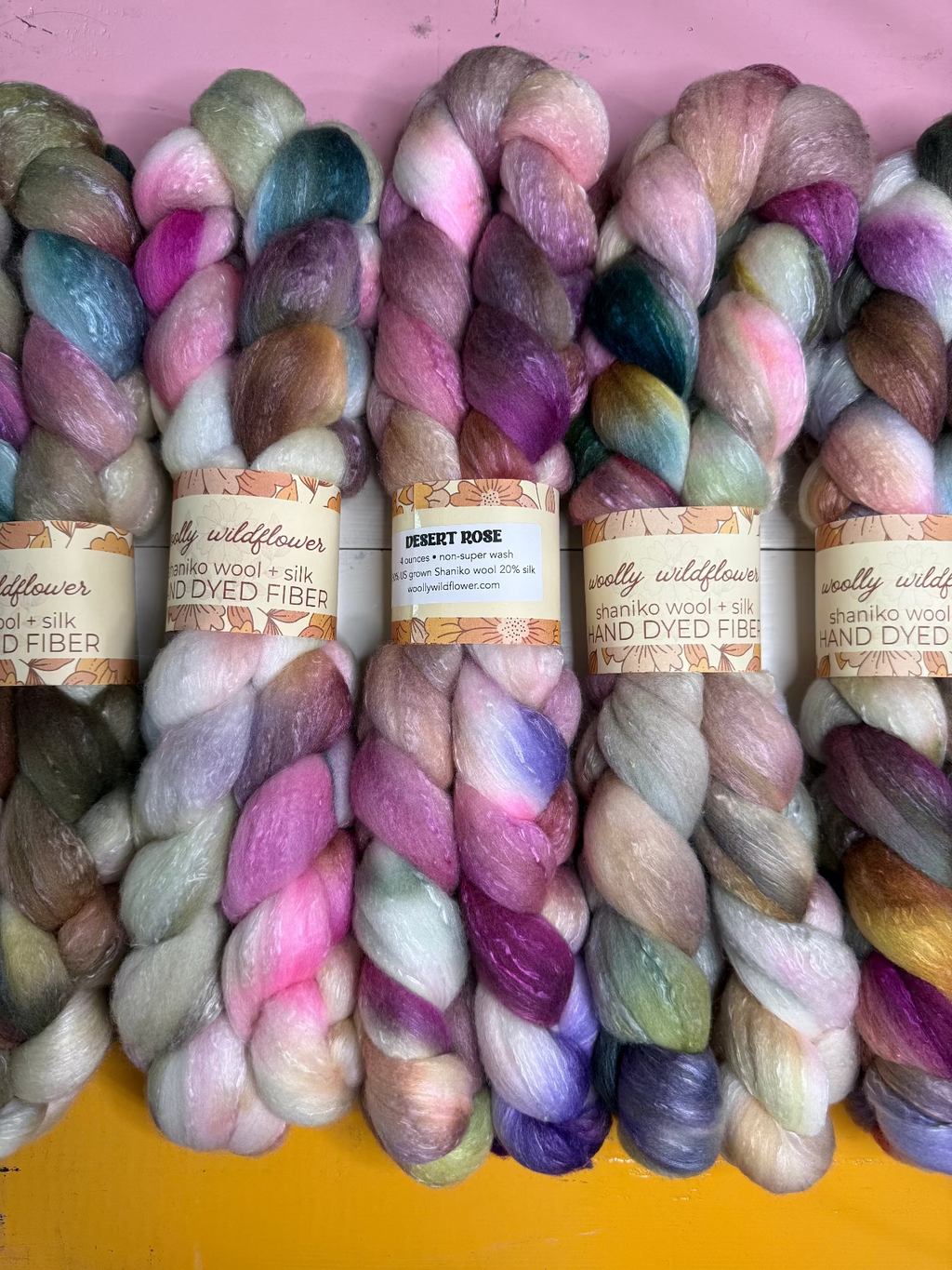 Five skeins of hand-dyed fiber yarn lined up side-by-side. The yarns feature a blend of pastel and vibrant colors including pink, purple, blue, green, and gold, with paper labels wrapped around the center of each skein.
