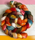 A coiled bundle of soft, twisted wool roving or fiber showing a multicolor mix of orange, red, blue, yellow, brown, pink, and cream, photographed against a painted background with pink, white, and yellow sections.