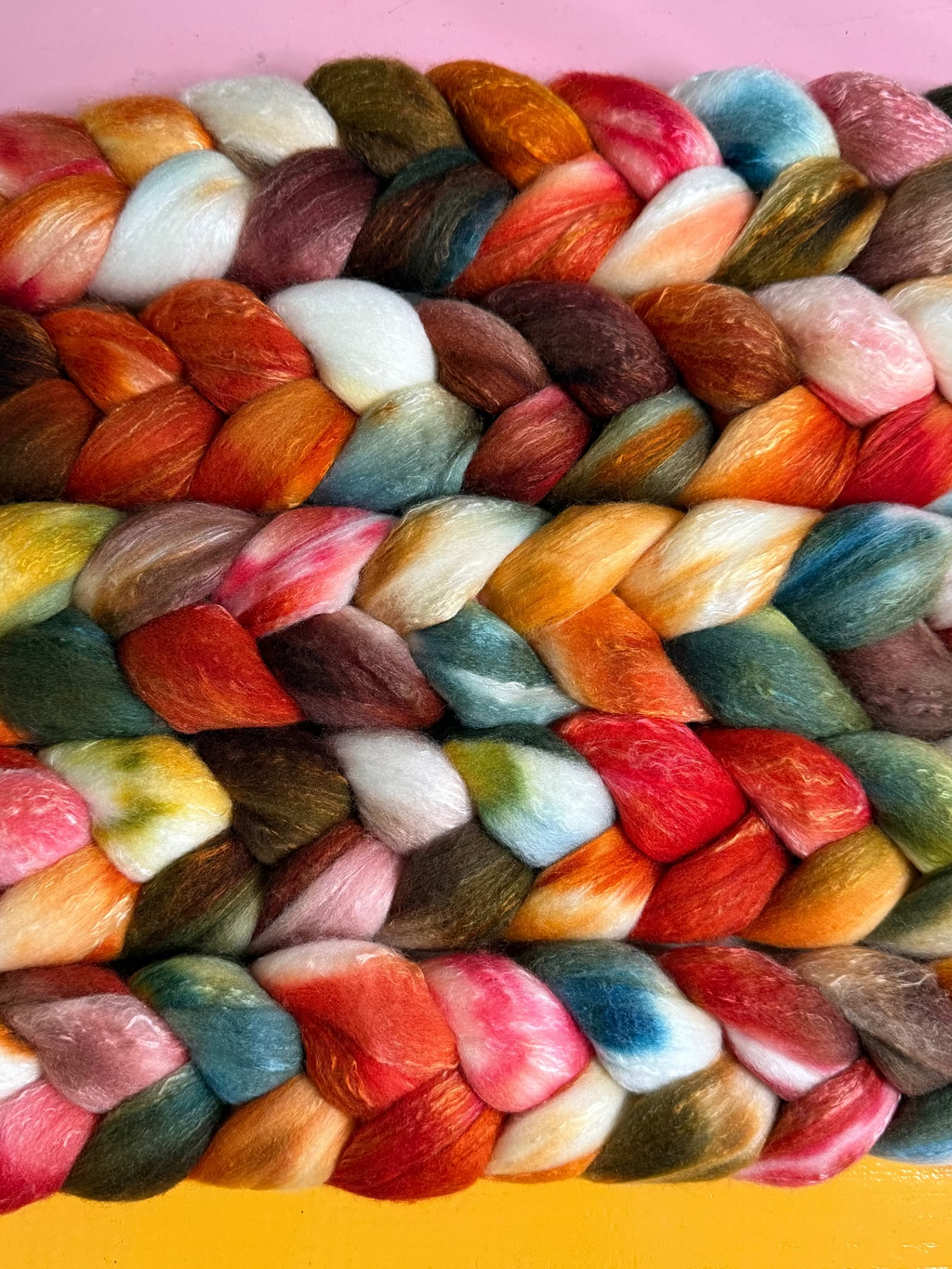 A close-up of braided wool roving or fiber bundles arranged in rows, showing a multicolor mix of orange, red, pink, blue, green, cream, and brown tones against a pink and yellow background.