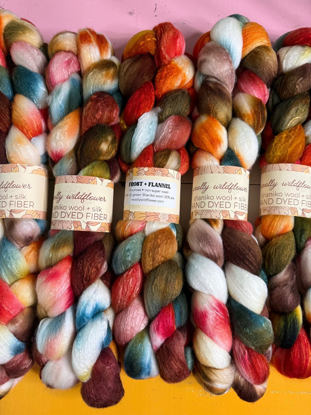 Six skeins of hand-dyed wool fibers arranged side-by-side against a pink and yellow background. Each skein has a paper label with the brand 'Wildflower'.