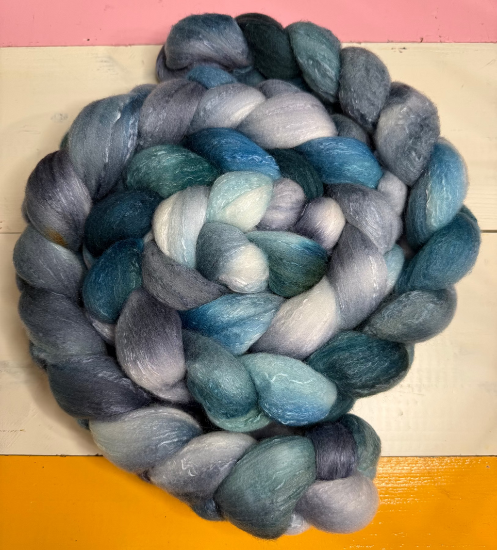 A coiled skein of fluffy, variegated wool roving in shades of blue, teal, gray, and white, photographed from above on a painted surface with pink, cream, and yellow sections.