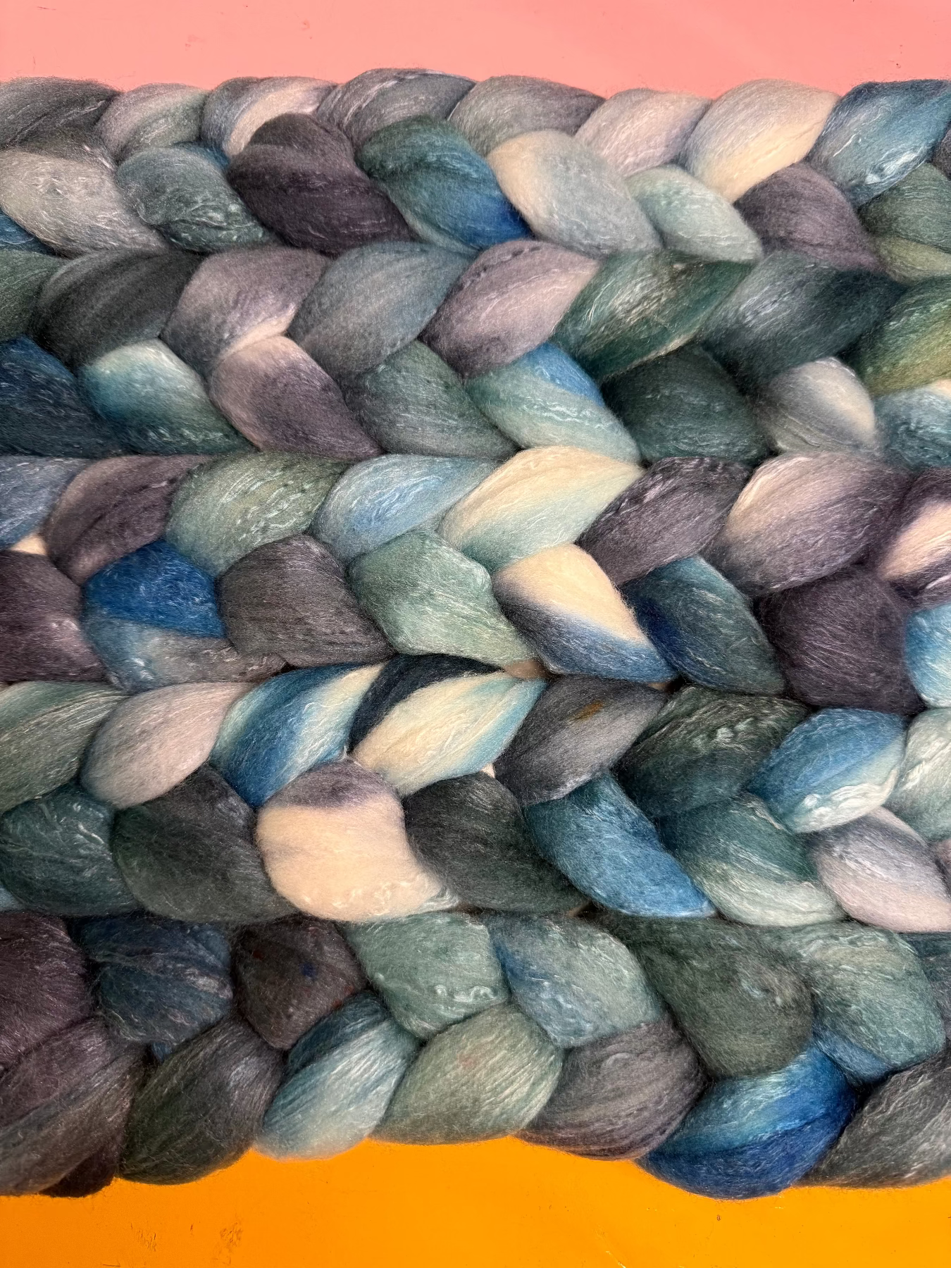 A close-up of a thick braided rope made from variegated fibers in shades of blue, teal, gray, and cream, photographed against a bright orange background.
