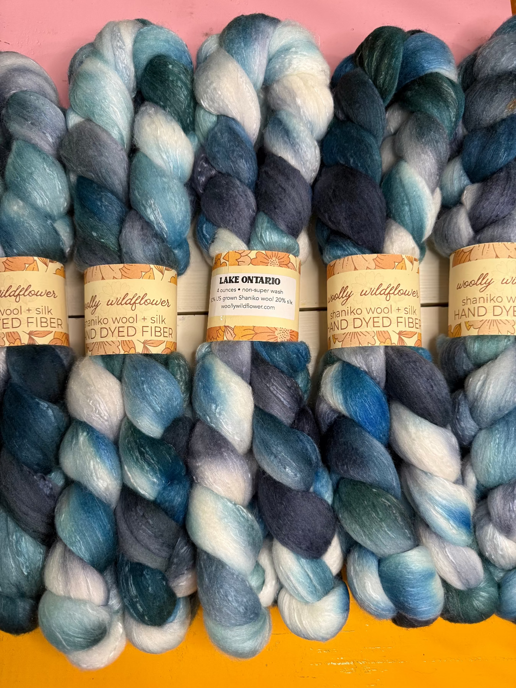 Five skeins of variegated hand-dyed wool roving in shades of blue, teal, navy, and white, arranged side-by-side with paper labels. The labels read 'Woolly Wildflower' and 'Lake Ontario'.