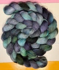 A coiled bundle of variegated wool roving or yarn in blended shades of teal, blue, purple, and green, photographed against a painted background with pink, white, and orange sections.