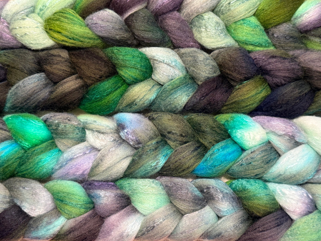 Close-up of braided yarn or fiber strands in a variegated mix of green, teal, purple, gray, brown, and cream tones.