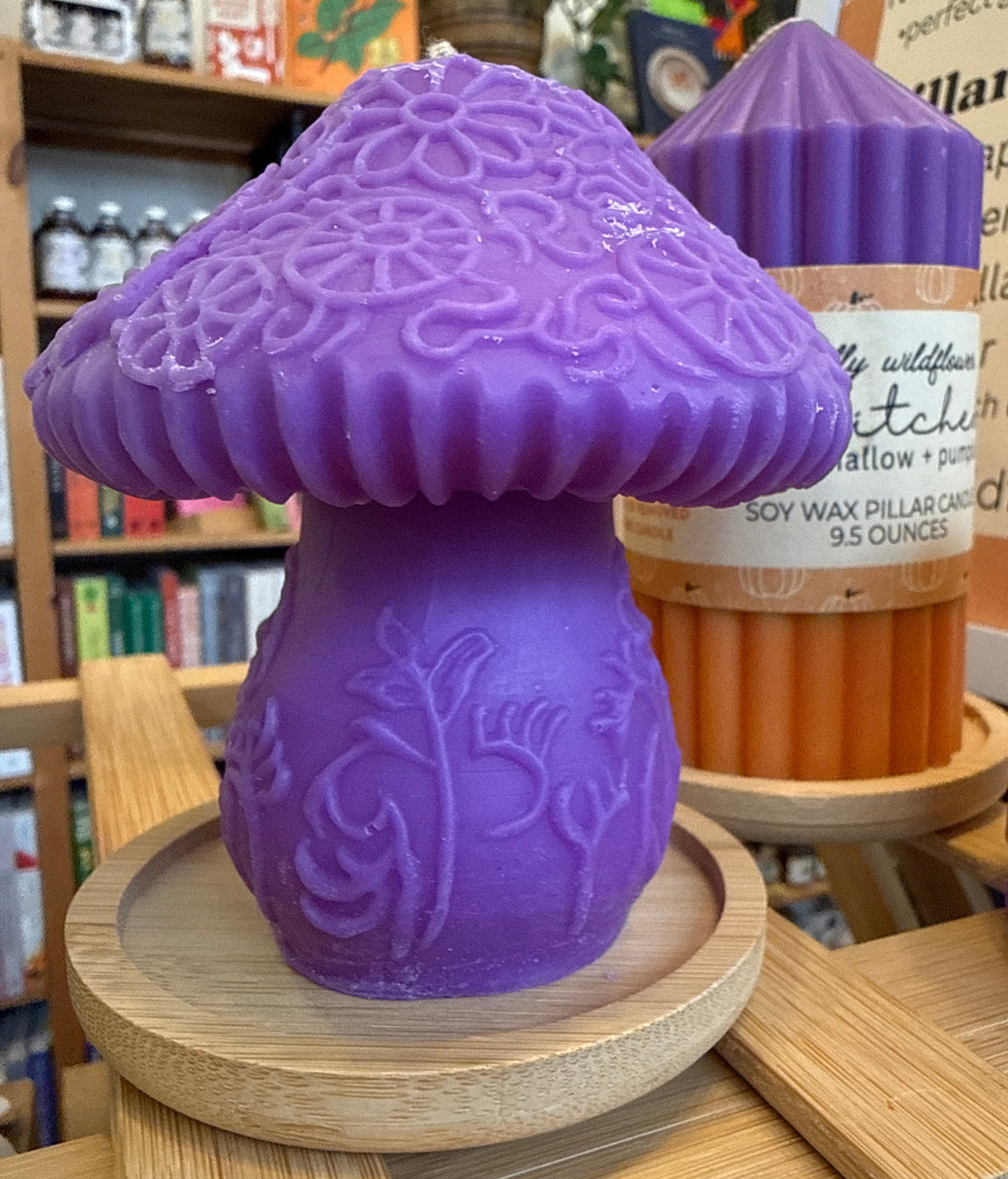 A purple mushroom-shaped soy wax candle on a round wooden base, displayed in a shop setting with other candles in the background.
