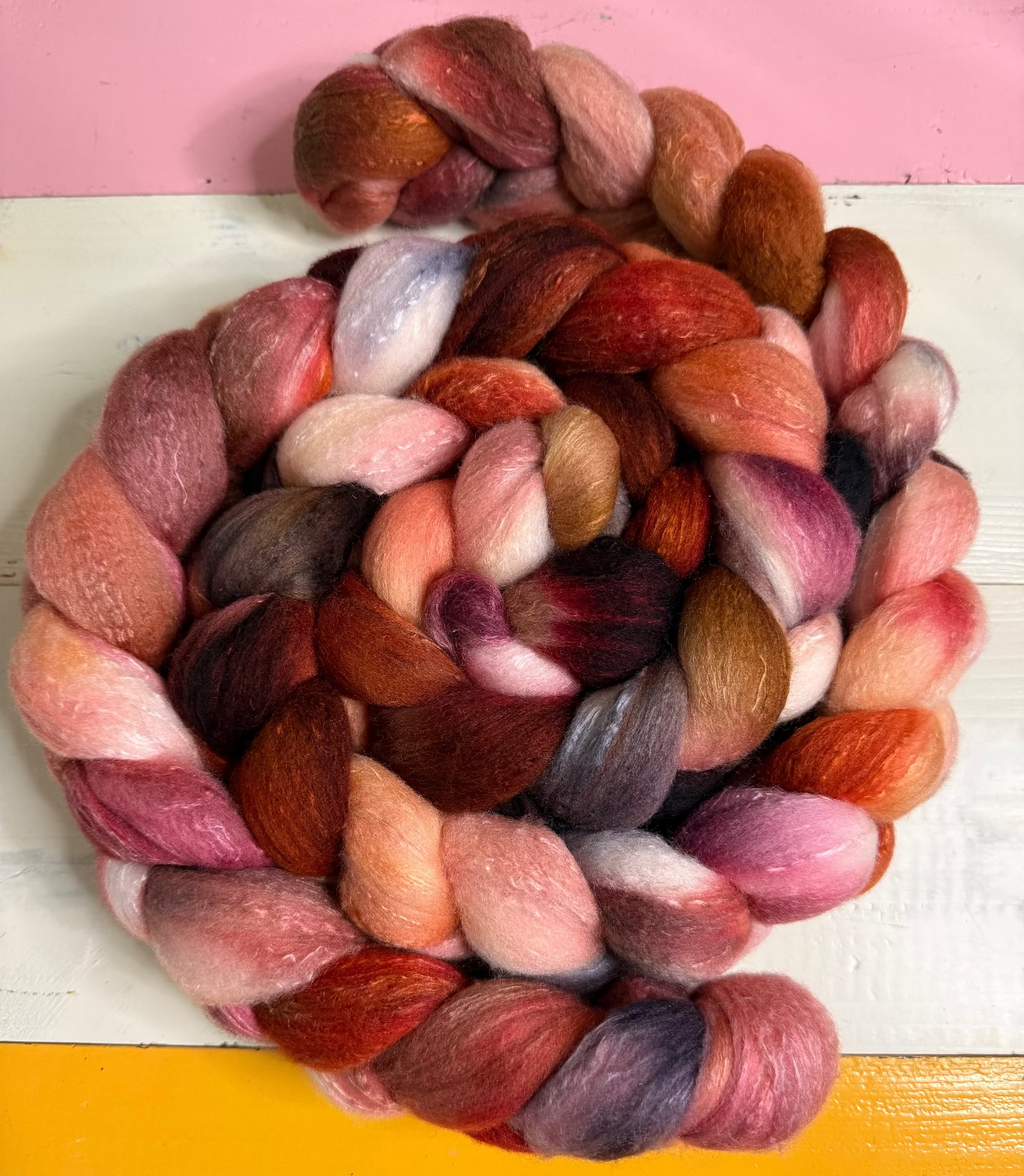 A coiled bundle of soft, fuzzy fiber roving in blended warm and cool tones including pink, red, orange, brown, purple, and cream, photographed against a painted background with pink, white, and yellow sections.