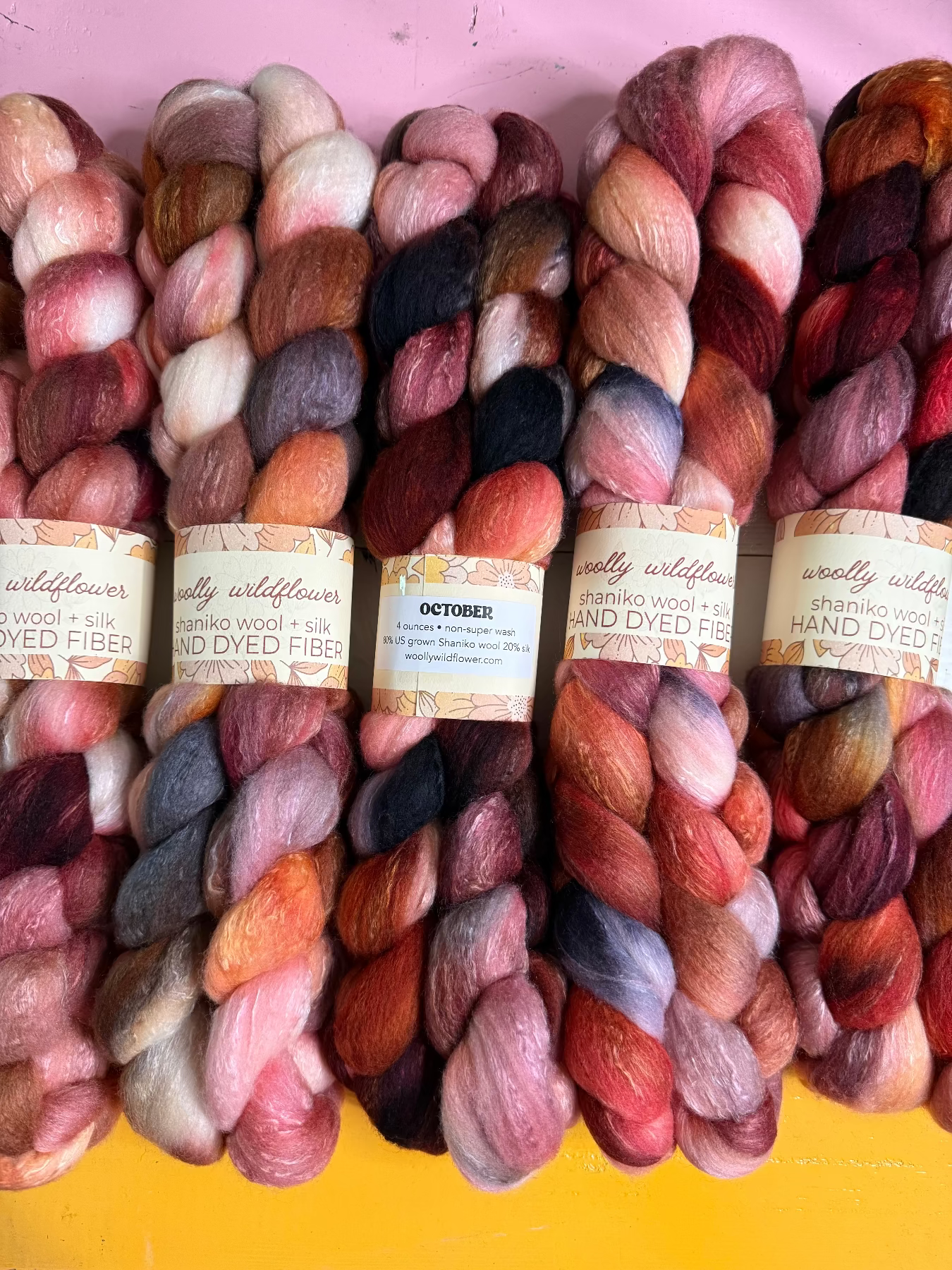 Six skeins of hand-dyed wool fiber yarn from Woolly Wildflower, arranged side-by-side. The yarns display a rich variety of colors including pink, red, orange, purple, and brown, with paper labels wrapped around the center skeins.