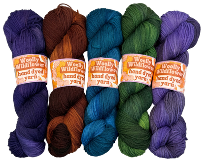 Five skeins of hand-dyed yarn in purple, brown, blue, green, and purple on a white background with 'Woolly Wildflowers' labels.