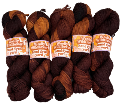 Five skeins of hand-dyed yarn in various brown shades on a white background with 'Woolly Wildflower' labels.